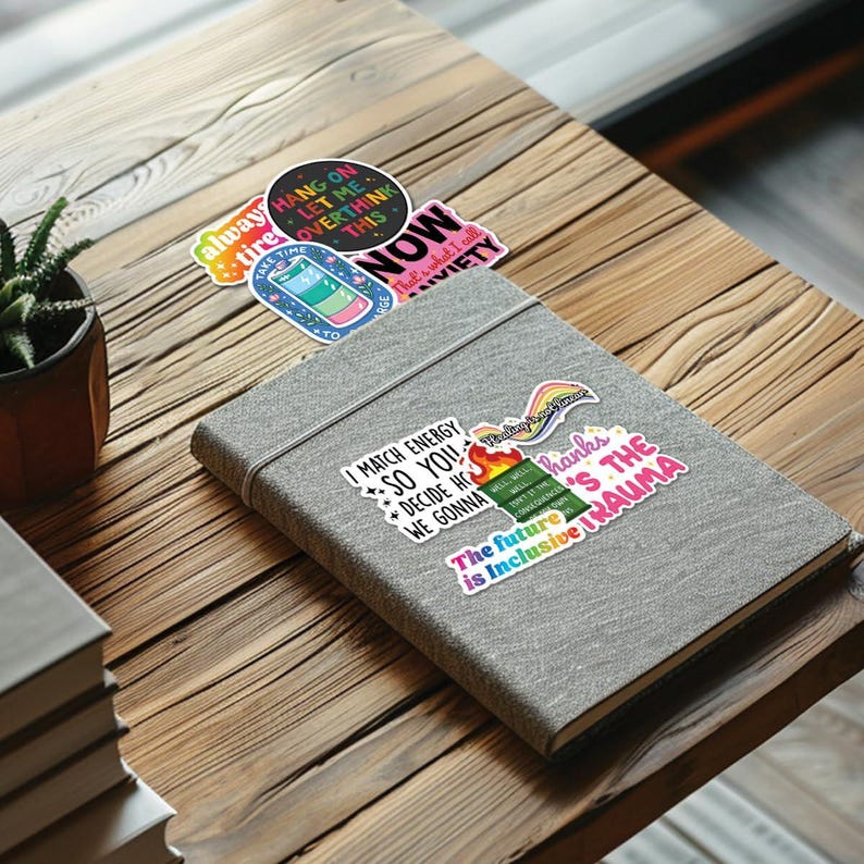 May include: A gray notebook with colorful stickers on the cover. The stickers feature phrases like "I match energy so you decide how we gonna" and "The future is inclusive trauma".