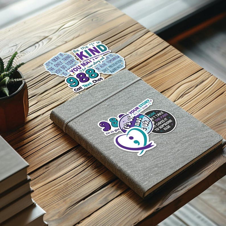 May include: A set of stickers with mental health awareness messages. The stickers are on a gray notebook. The stickers feature phrases like "It's okay if your story isn't over", "Be the "I" in kind", "Don't take the pain, it gives it to someone else", and "988 Call Text Chat".