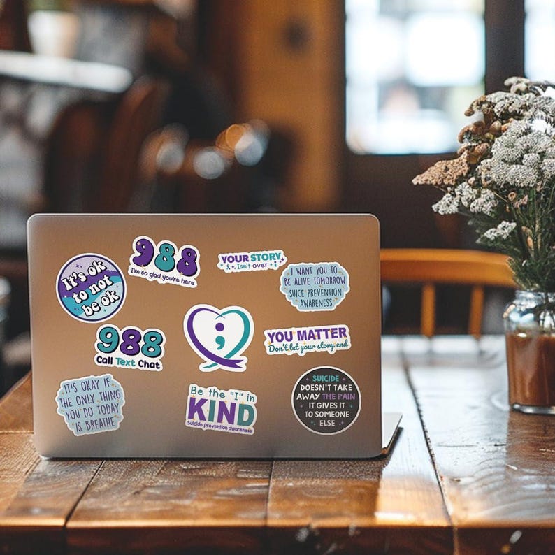May include: A silver laptop computer with several colorful stickers on the lid. The stickers feature messages of hope and support, including "It's ok to not be ok", "988 Call Text Chat", "Your Story Isn't Over", "Be the "I" in Kind", "You Matter", and "Suicide Doesn't Take Away the Pain".