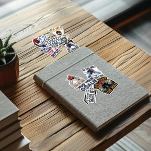 May include: A gray fabric-covered notebook with several hockey-themed stickers on a wooden table. The stickers feature phrases like "My goal is to deny" and "I'm the wall," along with hockey player illustrations. A small potted plant is visible.