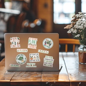 May include: A laptop computer with several stickers on it. The stickers feature illustrations of plants and text about plants. The text on the stickers includes: "Home is where my plants are", "Just one more plant I promise", "My plants are rooting for me", "Just a girl standing in front of her plants asking them not to die", "Pot head", "My plants and I talk shit about you", "Ask me about my plants".