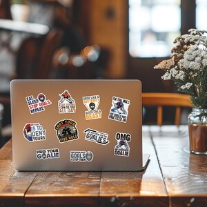 May include: A laptop covered in hockey-themed stickers. The stickers feature phrases like "Eat. Sleep. Stop Goals. Repeat." and "Shut Your Five Hole." Other stickers show hockey players and goalie-related sayings. The laptop sits on a wooden table.