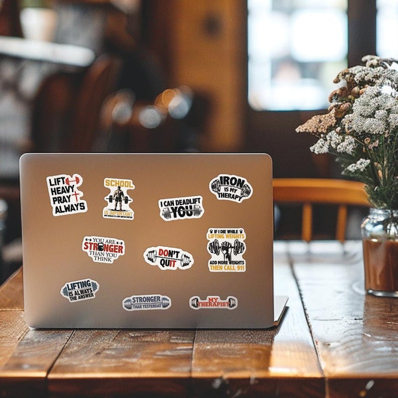 May include: A silver laptop computer with various colorful stickers featuring motivational quotes and images related to weightlifting. The stickers include phrases like "Lift Heavy Pray Always", "Iron is My Therapy", "I Can Deadlift You", and "You Are Stronger Than You Think".