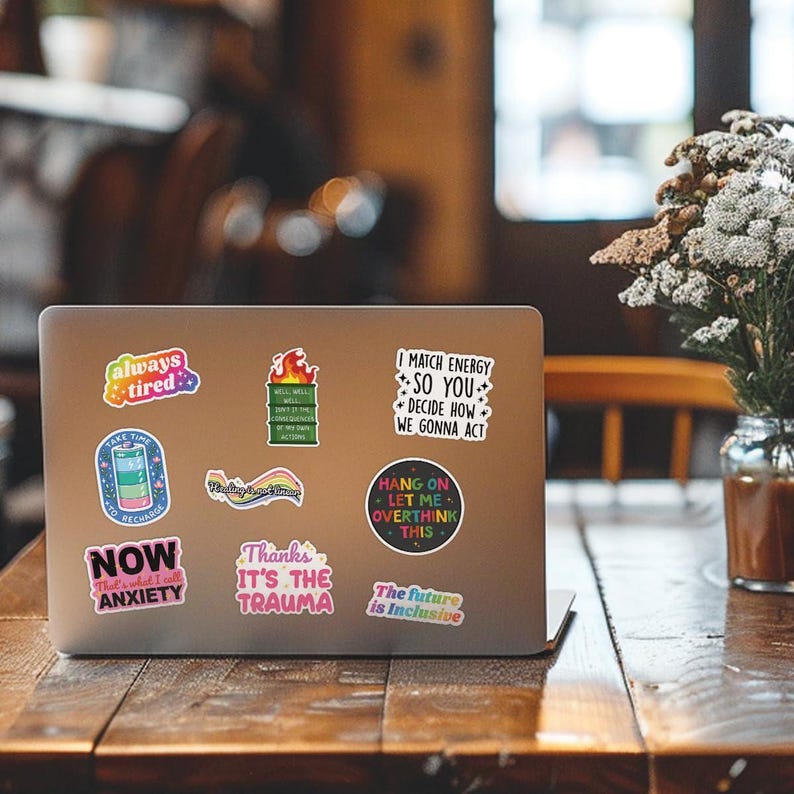 May include: A silver laptop with several colorful stickers on the lid. The stickers have various phrases on them, including "always tired", "take time to recharge", "now that's what I call anxiety", "thanks it's the trauma", "I match energy so you decide how we gonna act", "hang on let me overthink this", and "the future is inclusive".
