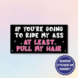May include: Black bumper sticker or magnet with white and pink text that reads "IF YOU'RE GOING TO RIDE MY ASS AT LEAST PULL MY HAIR." The sticker has a blue circular design that says "BUMPER STICKER OR MAGNET."