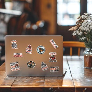 May include: A silver laptop computer with several stickers on the lid. The stickers feature various welding-related designs and sayings, such as "Flash Me I'm a Welder", "Welding Makes Me Happy", and "Yes, I Know I Am On Fire Let Me Finish This Weld".