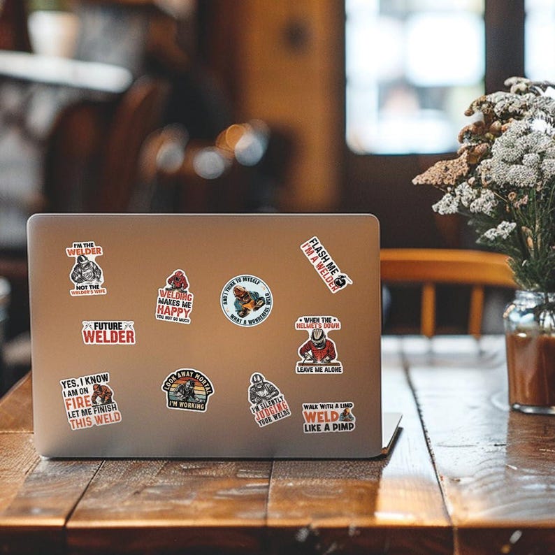 May include: A silver laptop computer with several stickers on the lid. The stickers feature various welding-related designs and sayings, such as "Flash Me I'm a Welder", "Welding Makes Me Happy", and "Yes, I Know I Am On Fire Let Me Finish This Weld".