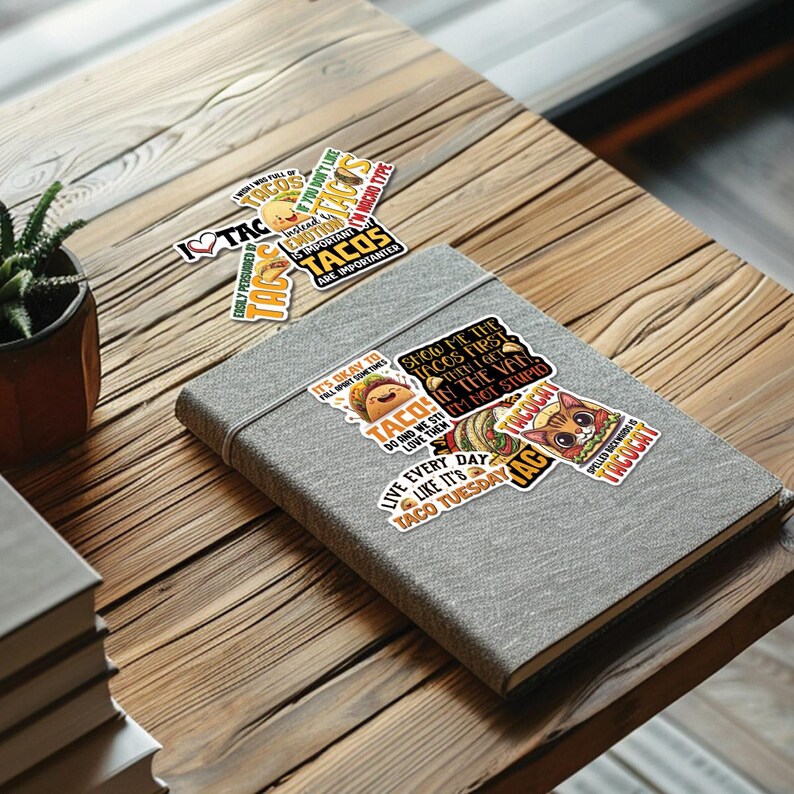 K&ouml;nnte beinhalten: Eine Sammlung farbenfroher Aufkleber zum Thema Tacos auf einem grauen Notizbuch. Die Aufkleber zeigen verschiedene Designs, darunter eine Katze, Tacos und Texte wie "Ich liebe Tacos" und "Tacos sind wichtig". Das Notizbuch liegt auf einer Holzoberfl&auml;che.