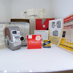 May include: A vintage Keystone movie camera with a brown and gray body, silver lens, and a handle. The camera is surrounded by vintage film boxes, instruction manuals, and a Sears film box. The items are arranged on a white surface.