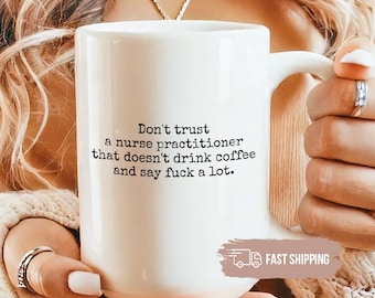 May include: White ceramic mug with a handle, featuring the text: "Don't trust a nurse practitioner that doesn't drink coffee and say fuck a lot." The mug is held by a person wearing rings and a beige sweater.