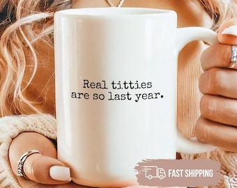 May include: A white ceramic mug featuring the black printed text 'Real titties are so last year.' The mug has a standard handle and a smooth finish, held in place to display the message clearly against a neutral background.