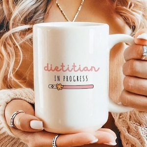 May include: A white ceramic mug with the words "dietitian IN PROGRESS" in pink lettering. A pink bar with a flower graphic is below the text. The mug has a large handle and is held by a person.