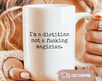 May include: A white ceramic mug featuring the black printed text, I'm a dietitian not a fucking magician. The mug has a standard cylindrical shape with a curved handle on the right side, designed for holding hot beverages in a comfortable manner.