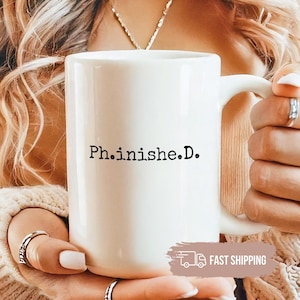 May include: A white ceramic mug with the text "Ph.inish.e.D." in black. The mug has a large handle and is held by a person wearing rings and a beige sweater.