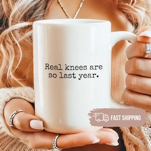 May include: White ceramic mug with the text "Real knees are so last year." The mug has a large handle and is held by a person. The mug is a standard size for coffee or tea.