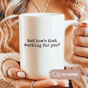 May include: White ceramic mug with the text "And how's that working for you?" in black. The mug has a large handle. The mug is held by a person. The mug is a standard size for tea or coffee.