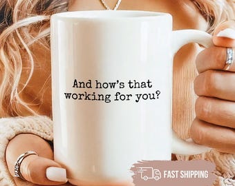 May include: White ceramic mug with the text "And how's that working for you?" in black. The mug has a large handle and is held by a person. The mug is a standard size for coffee or tea.