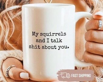 May include: A white ceramic mug featuring the black printed text My squirrels and I talk shit about you. The mug has a standard handle and a smooth finish, held in place with both hands. A small rectangular graphic with the text FAST SHIPPING is visible.