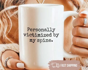 May include: White ceramic mug with the text "Personally victimized by my spine." The mug has a large handle and is held by a person wearing a beige sweater. The mug is a standard size for coffee or tea.