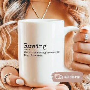 May include: A white ceramic mug with the word "Rowing" and its definition printed in black. The mug has a large handle and is held by a person. The mug is a standard size.