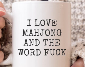 May include: White ceramic mug with the text "I LOVE MAHJONG AND THE WORD FUCK" in black, held by a person wearing a gray sweater. The mug is a standard size and shape, suitable for hot beverages.