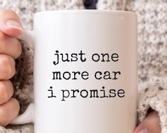 May include: A white ceramic mug featuring the black typewriter-style text "just one more car i promise" printed on the front. The mug has a standard handle and a smooth, glossy finish, designed for holding hot or cold beverages.