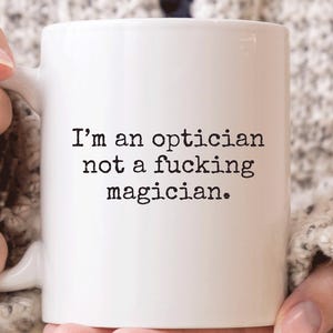 May include: White ceramic mug with the text "I'm an optician not a fucking magician." The mug is held by a person wearing a light grey jumper. The mug has a standard handle.