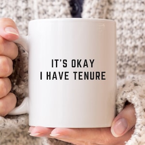 Könnte beinhalten: Weiße Keramik-Kaffeetasse mit schwarzem Text, der "Es ist okay, ich habe eine feste Anstellung" lautet.