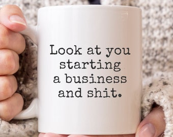 May include: White ceramic mug with the text "Look at you starting a business and shit." The mug is held by a person wearing a gray knit sweater. The mug is a standard size.