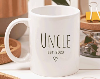 May include: White ceramic mug with a black inscription that reads "Uncle Est. 2023" with a small heart symbol. The mug is sitting on a white surface with a plant, a cork coaster, and a jar of paper clips in the background.
