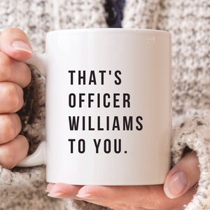 May include: White ceramic mug with the text "THAT'S OFFICER WILLIAMS TO YOU." in black. The mug is held by a person wearing a grey knit sweater. The mug is a standard size for coffee or tea.