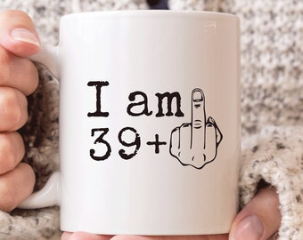 May include: White ceramic coffee mug with black text that reads "I am 39+" and a black and white illustration of a middle finger.