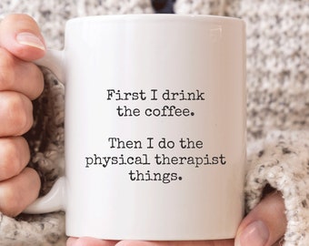 May include: A white ceramic mug with black text that reads, "First I drink the coffee. Then I do the physical therapist things." The mug is held by a person wearing a light gray sweater.