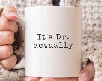 May include: White ceramic coffee mug with black text that reads "It's Dr. actually".