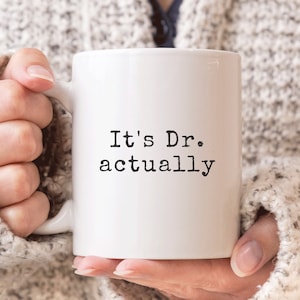 May include: White ceramic coffee mug with black text that reads "It's Dr. actually".