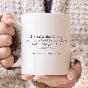May include: White ceramic mug featuring the quote "I would challenge you to a battle of wits, but I see you are unarmed." by William Shakespeare. The mug is held by a person wearing a cream-coloured sweater.