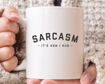 May include: White ceramic coffee mug with black text that reads "SARCASM - IT'S HOW I HUG -" The mug is being held by a person wearing a beige sweater.