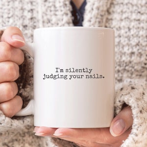 May include: White ceramic coffee mug with black text that reads "I'm silently judging your nails."