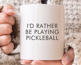 May include: White ceramic coffee mug with black text that reads "I'd rather be playing pickleball".