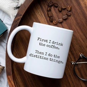 May include: White coffee mug with black text that reads "First I drink the coffee. Then I do the dietitian things."