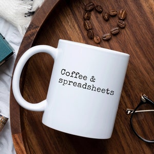 May include: A white ceramic mug with the text "Coffee & spreadsheets" sits on a wooden tray. Coffee beans are arranged in a heart shape. A vintage teal camera, glasses, and a potted plant are also on the tray.