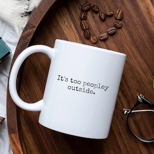 以下が含まれることがあります： 黒い文字で「It's too peopley outside.」と書かれた白いセラミック製のコーヒーカップ