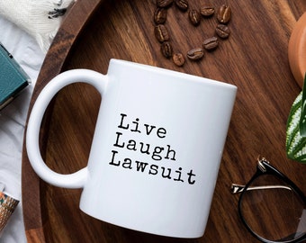 May include: White coffee mug with black text that reads "Live Laugh Lawsuit". The mug is on a wooden tray with coffee beans and a green plant.