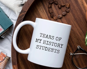 May include: White coffee mug with black text that reads "Tears of My History Students". The mug is on a wooden tray with coffee beans, a plant, and a pair of glasses.