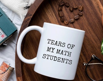 May include: White ceramic coffee mug with black text that reads "Tears of My Math Students". The mug is on a wooden tray with coffee beans, a plant, and a camera.