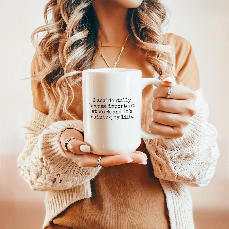 May include: White ceramic mug with the text "I accidentally became important at work and it's ruining my life." The mug has a large handle and is held by a person. The mug is a standard size for coffee or tea.