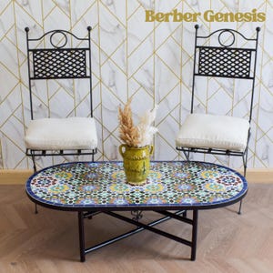 May include: A black metal oval table with a colorful mosaic tile top. Two black metal chairs with white cushions are placed on either side of the table. The table and chairs are in front of a white wall with a gold geometric pattern.