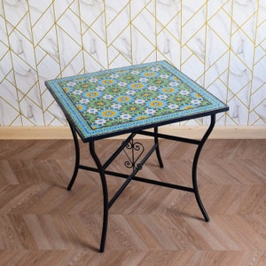 May include: A small square table with a black metal frame and a colourful mosaic tile top. The tile design features a repeating pattern of blue, green and yellow flowers.