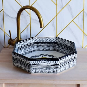 Op de afbeelding: Achtekantige keramische wastafel met een geometrisch patroon in zwart en wit. De wastafel heeft een geel en groen bloemmotief in het midden.