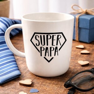Peut inclure: Mug en céramique blanche avec l'inscription "SUPER PAPA" dans un losange noir. La tasse a une anse incurvée et repose sur une surface en bois. Des cœurs en bois décoratifs et une paire de lunettes noires sont également visibles.